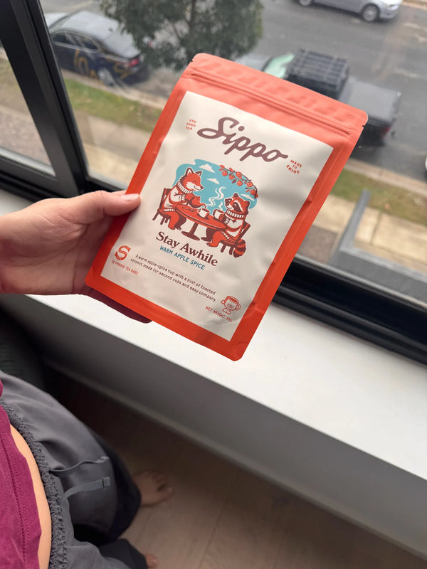 A hand holds the Stay Awhile apple spice tea pack against an overcast street view